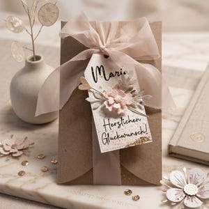 May include: A brown paper envelope adorned with a sheer light pink ribbon bow and a white rectangular tag. The tag features the name Maria, the text Herzlichen Gl&uuml;ckwunsch!, and a decorative arrangement of small pink flowers and gold accents.
