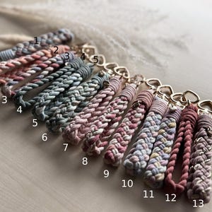 May include: Thirteen braided cord keychains with gold-toned heart-shaped clasps, numbered 1 through 13. The keychains feature various color combinations including shades of pink, blue, grey, and cream, with some strands interwoven with metallic thread.