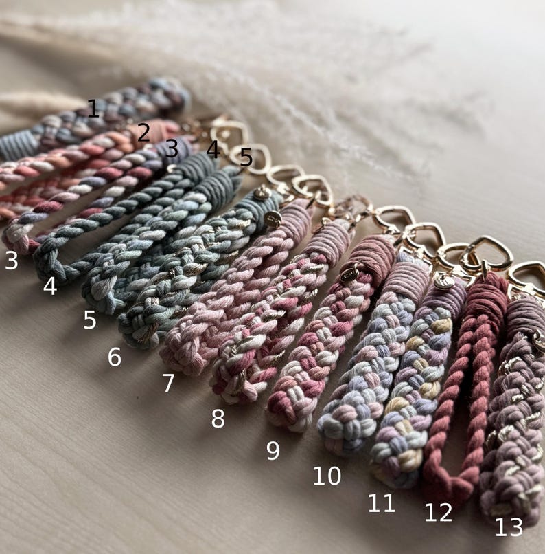 May include: Thirteen braided cord keychains with gold-toned heart-shaped clasps, numbered 1 through 13. The keychains feature various color combinations including shades of pink, blue, grey, and cream, with some strands interwoven with metallic thread.