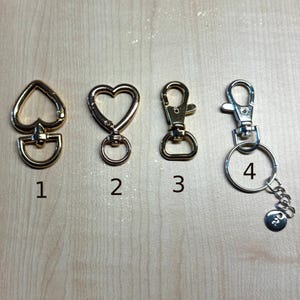 May include: Four metal swivel clasps labeled one through four. Number one is a gold heart-shaped clasp with a D-ring. Number two is a gold heart-shaped clasp with a circular ring. Number three is a gold lobster claw clasp. Number four is a silver lobster claw clasp with a split ring and a small circular charm.