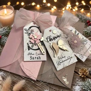 May include: Two rectangular gift tags with pink ribbon bows. One tag features a large black letter S, a pink flower, and the text "Alles Liebe Sarah" and "VIEL GL&Uuml;CK". The other tag displays gold angel wings and the text "Frohe Weihnachten".