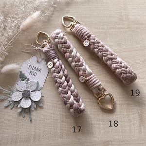 May include: Three braided macrame wristlets in shades of mauve, pink, and cream, each featuring gold-tone heart-shaped clasps and small circular letter charms. A white tag with the text THANK YOU and a paper flower decoration are attached.