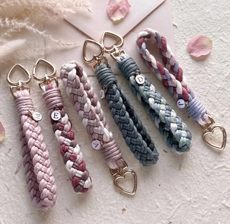 May include: Six braided cord keychains with gold-tone heart-shaped clasps. The keychains feature various color combinations including solid pink, multi-toned pink, grey, and blue. Several keychains include small circular charms with letters M, E, A, S, D, and J.