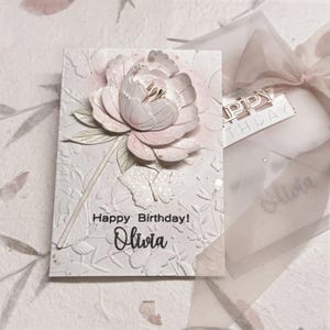 May include: A rectangular greeting card featuring a three-dimensional pale pink peony flower, embossed butterfly and floral patterns, and the printed text Happy Birthday! Olivia. The card is paired with a translucent envelope tied with a ribbon.