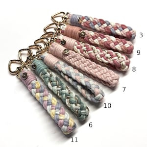May include: A collection of six braided fabric keychains with gold-toned heart-shaped clasps and small circular metal tags. Each keychain features a unique color combination of pastel threads, some interwoven with metallic gold strands, labeled 3, 9, 8, 7, 10, 6, and 11.