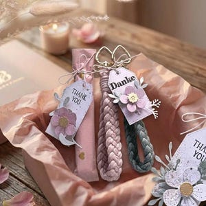 May include: Two braided wristlet keychains, one light pink and one dark gray, each featuring a gold-tone heart-shaped clasp and a rectangular tag. The tags are decorated with paper flowers and the word Danke or the phrase Thank You in black text.