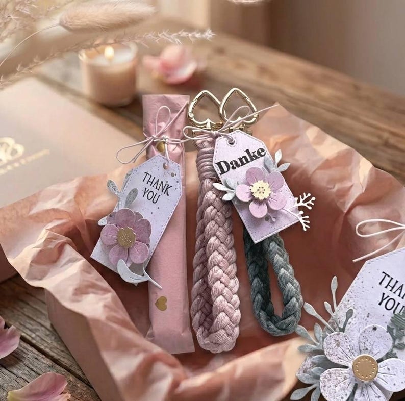 May include: Two braided wristlet keychains, one light pink and one dark gray, each featuring a gold-tone heart-shaped clasp and a rectangular tag. The tags are decorated with paper flowers and the word Danke or the phrase Thank You in black text.
