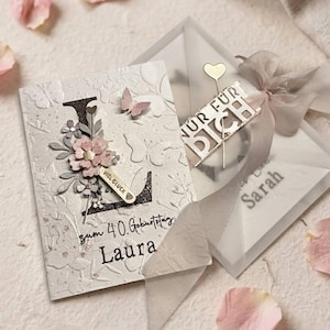May include: A white greeting card with a large black letter L, floral embellishments, a small pink butterfly, and the text "zum 40. Geburtstag Laura" and "Viel Glück". Beside it, a translucent envelope with a ribbon and "Nur für dich" text.