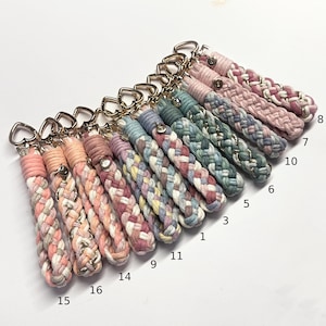 May include: A collection of braided cord keychains featuring gold-tone heart-shaped clasps. Each keychain is crafted from multi-colored cotton rope in various pastel and muted tones, with some strands interwoven with metallic gold thread.