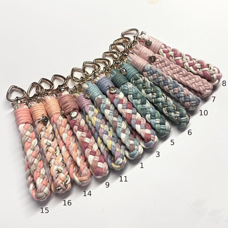 May include: A collection of braided cord keychains featuring gold-tone heart-shaped clasps. Each keychain is crafted from multi-colored cotton rope in various pastel and muted tones, with some strands interwoven with metallic gold thread.