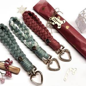 May include: Three braided macrame wristlets with gold-toned heart-shaped lobster clasps. One is solid dark green, one is a variegated mix of sage green and white, and one is solid deep red. Each features a small circular metal charm attached.