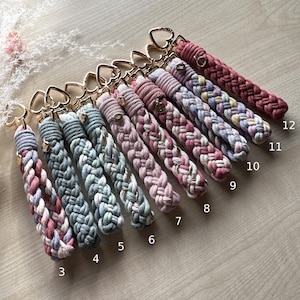 May include: A collection of twelve braided macrame keychains with gold-toned heart-shaped clasps, numbered three through twelve. Each keychain features a unique color combination of cotton cord, including shades of blue, pink, mauve, and cream.