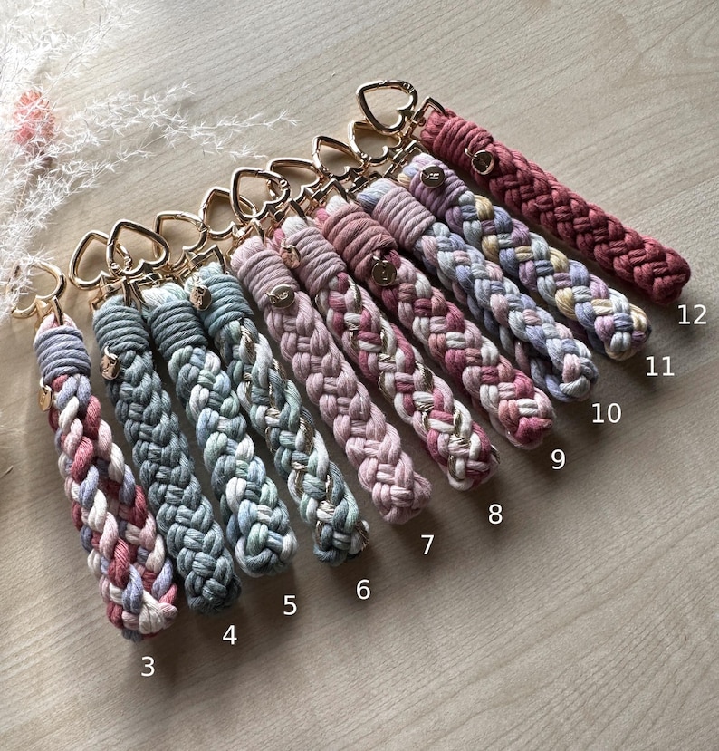 May include: A collection of twelve braided macrame keychains with gold-toned heart-shaped clasps, numbered three through twelve. Each keychain features a unique color combination of cotton cord, including shades of blue, pink, mauve, and cream.