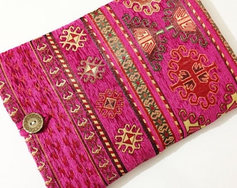 Pode incluir: Uma capa para laptop de tecido rosa com um padrão tradicional de kilim turco. A capa tem um fecho de botão castanho.
