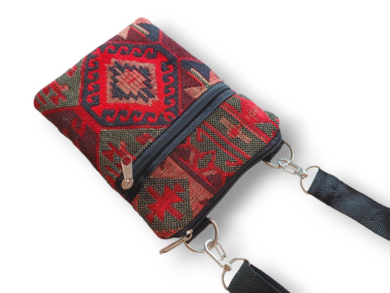 May include: A small rectangular crossbody bag featuring a woven fabric with a geometric pattern in shades of red, black, and dark green. The bag includes a black zippered closure, silver-toned metal hardware, and a black adjustable fabric strap.