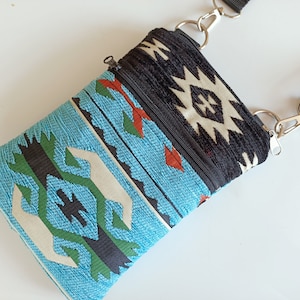 May include: A rectangular crossbody bag featuring a woven fabric with a geometric pattern in turquoise, black, white, and orange. The bag includes a black zippered top closure, a black zippered front pocket, and a black adjustable nylon strap.