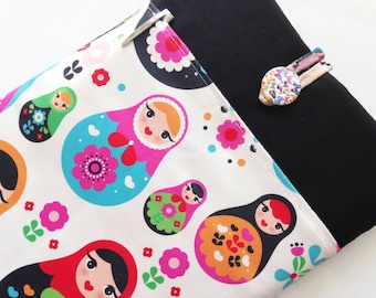Pode incluir: Uma bolsa de tecido branco com uma aba de tecido preto. A bolsa é decorada com um padrão colorido de bonecas russas aninhadas, flores e corações. A aba tem um botão decorativo com um padrão floral.