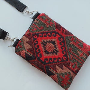 May include: A rectangular crossbody bag featuring a woven fabric with a geometric pattern in shades of red, black, and tan. The bag includes a black nylon strap attached with silver-toned metal swivel clips and rings at the top corners.