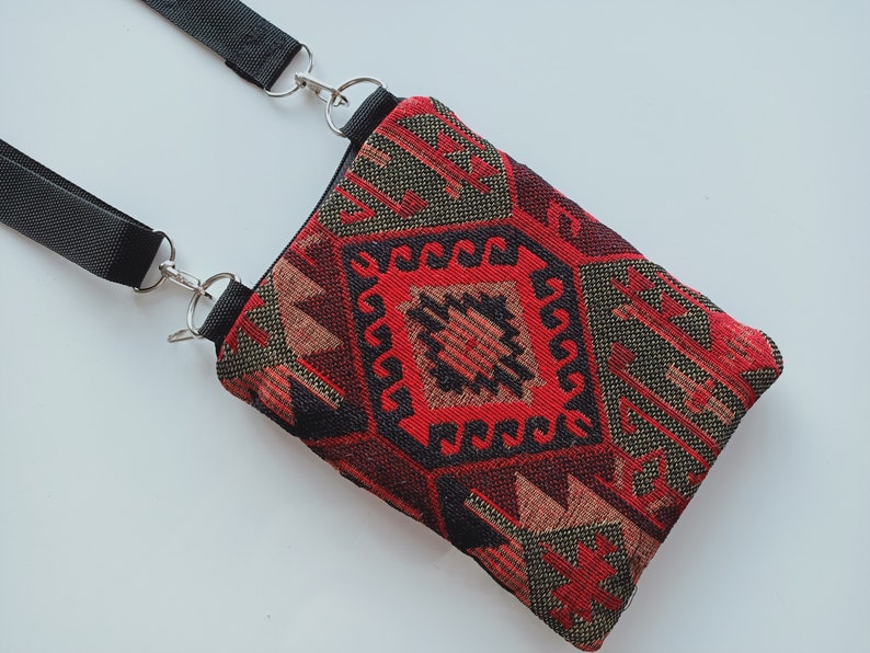 May include: A rectangular crossbody bag featuring a woven fabric with a geometric pattern in shades of red, black, and tan. The bag includes a black nylon strap attached with silver-toned metal swivel clips and rings at the top corners.
