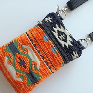 May include: A small crossbody bag featuring a textured fabric with a geometric pattern in orange, black, green, and cream. The bag includes a black zippered top closure, silver-toned metal hardware, and an adjustable black nylon shoulder strap.