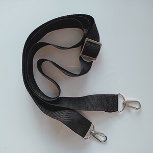 May include: A black adjustable nylon strap featuring a silver-toned metal slider for length adjustment and two silver-toned metal swivel lobster claw clasps attached to each end, designed for use as a replacement shoulder strap for bags.