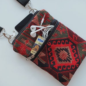 May include: A small rectangular crossbody bag featuring a woven red, black, and tan geometric pattern. The bag has a black adjustable strap, a main zippered compartment containing a black smartphone, and a front zippered pocket holding white wired earbuds and a Visa credit card.