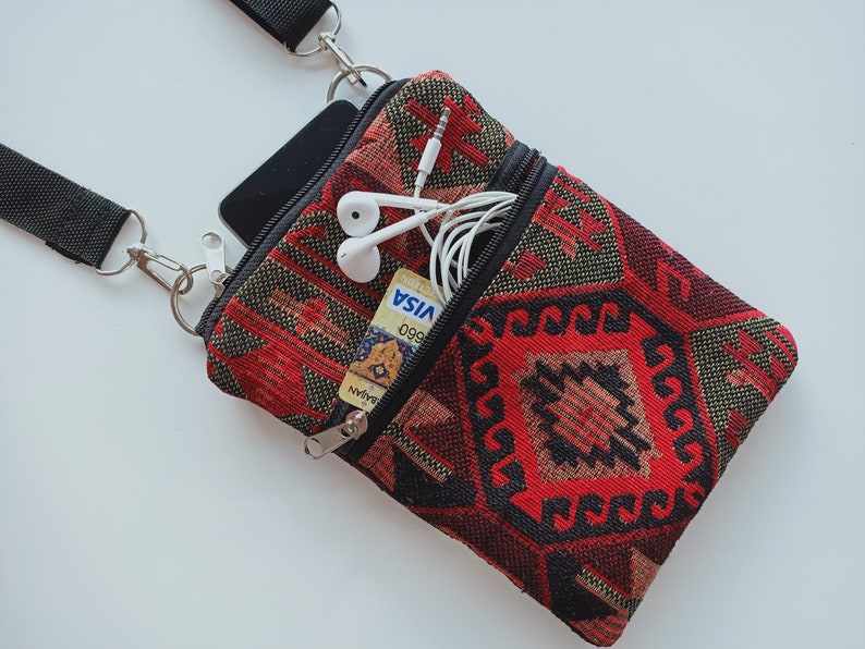May include: A small rectangular crossbody bag featuring a woven red, black, and tan geometric pattern. The bag has a black adjustable strap, a main zippered compartment containing a black smartphone, and a front zippered pocket holding white wired earbuds and a Visa credit card.