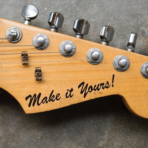 Può includere: Primo piano della paletta di una chitarra elettrica vintage con sei meccaniche. La chitarra è in legno e ha una finitura naturale. La paletta ha un adesivo nero che dice "Make it Yours!"
