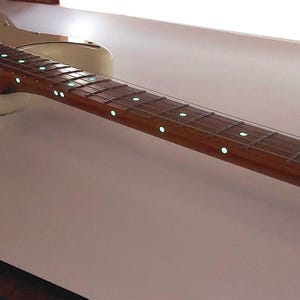 May include: Close-up of a guitar fretboard with glow-in-the-dark dot markers. The fretboard is made of wood and has a natural finish. The dots are evenly spaced and are a bright green colour.