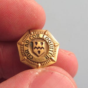 May include: A small, gold-colored pin with an octagonal shape. The pin features a crest design with the words "AD COR LOQUITUR" around the edge. The crest has a shield with a design inside. The pin is held between two fingers.