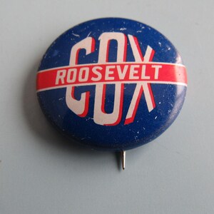May include: A vintage blue and white political campaign button for Cox and Roosevelt. The button features the names in white lettering on a blue background, with a red stripe. A metal pin is attached to the back.