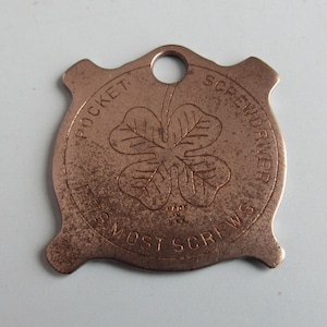 May include: A copper-colored pocket screwdriver with a four-leaf clover design in the center. The text "POCKET SCREWDRIVER" and "MOST SCREWS" are etched around the edge. The tool has a small hole at the top.