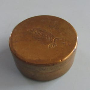 May include: A small, round, copper-colored container with a lid. The lid has the word "Lavador" engraved on it. The container is sitting on a white surface. The container is made of metal.
