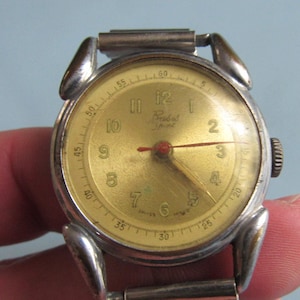 May include: A vintage wristwatch with a round silver-tone metal case and a metallic gold-colored dial. The face features black Arabic numerals, a red second hand, and the text "Pobeda" printed in black. It includes a flexible metal link band.