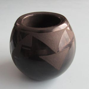 May include: A dark brown, rounded vase with a wide opening. The vase features a geometric design of triangles and other shapes carved into the surface. The vase is sitting on a light blue surface.