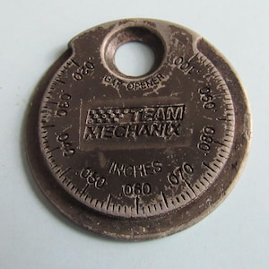 May include: A silver-colored spark plug gap gauge with a circular design. The gauge has a hole at the top and includes measurements in inches, ranging from .020 to .100. The text "TEAM MECHANIX" is visible.