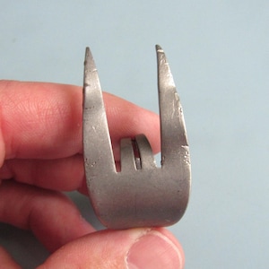 May include: A silver-toned metal ring crafted from a repurposed fork, featuring two prominent, upward-curving tines that extend from the band. The metal surface displays a matte, weathered texture with visible signs of wear and industrial shaping.