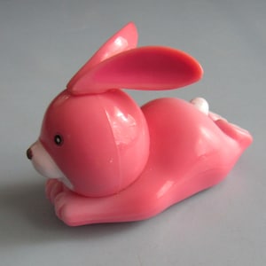 May include: A small, smooth, pink plastic rabbit figurine in a reclining position. The rabbit has long, upright ears, a white muzzle area, a small brown nose, and a tiny white tail visible at the rear. The surface of the toy is glossy.