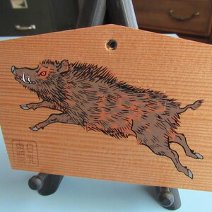 May include: A wooden plaque features a hand-painted image of a boar, predominantly brown and black, with reddish eyes and white tusks. The boar is depicted mid-leap, its body angled to the left.  The plaque's light brown wood grain is visible.  Japanese characters are stamped in a rectangular box in the upper right corner and a smaller rectangular box in the lower left corner.  The plaque is displayed on a simple black stand.