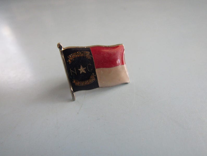May include: A small, enameled pin in the shape of the North Carolina state flag. The pin features a black section with gold lettering and a star, a red section, and a tan section. The pin has a gold-colored border and a small flag pole.