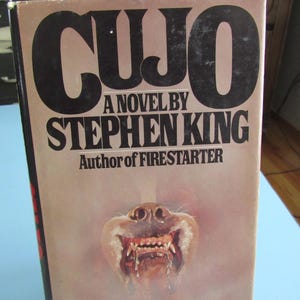 May include: Vintage paperback book titled "CUJO" by Stephen King. The cover features the title in large black letters, the author's name, and a menacing illustration of a dog's snarling face with exposed teeth and saliva.