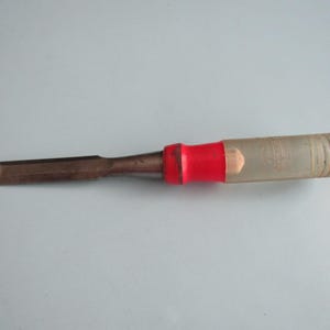 May include: A woodworking chisel with a metal blade and a clear plastic handle. The handle has a red band near the blade. The chisel is designed for shaping and cutting wood. The blade is approximately 1 inch wide.
