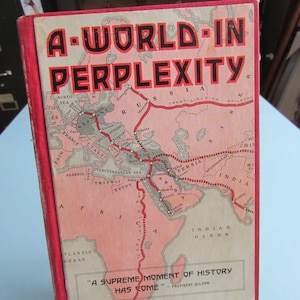 May include: A red book titled "A-World-In Perplexity" with a map of the world on the cover. The map shows a red line tracing a path across the globe. The text "A Supreme Moment of History Has Come" is printed below the map, attributed to President Wilson.