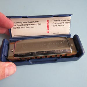 May include: A silver Hohner Blues Harp harmonica with ten numbered holes, housed in a blue plastic case. The case lid features an instructional insert with the text Hohner MS-System Component Replacement Instructions and German text.