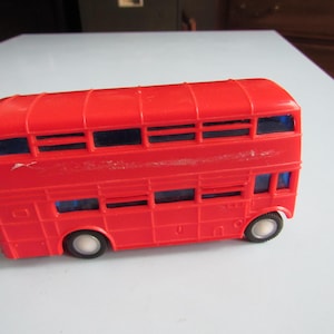 May include: A bright red plastic toy double-decker bus featuring two levels of windows with dark blue tinted plastic inserts. The bus has a molded exterior with visible panel lines, a roof with raised ridges, and four small black wheels with white centers.