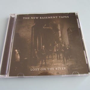 May include: A CD case featuring the album "Lost on the River" by The New Basement Tapes. The cover art is a sepia-toned photograph of a group of people standing in a narrow alleyway. The album title and artist name are printed in white.