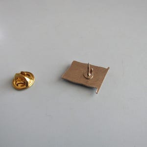 May include: A gold-colored flag pin with a pin back and a separate gold-colored clutch. The flag pin is a rectangular shape with a small pin extending from the back. The clutch is round.