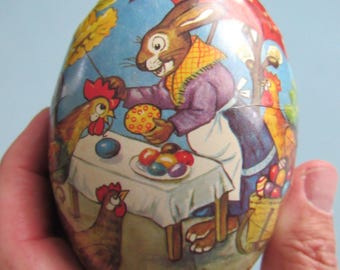 May include: An egg-shaped Easter decoration featuring a colorful illustration of a rabbit selling eggs and treats to chickens. The egg has a red umbrella and a table with painted eggs. The overall theme is Easter.