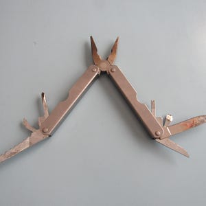 May include: A silver multi-tool with pliers, a saw, a knife, and other tools. The multi-tool is open, displaying its various functions. The tools are made of metal and are on a light blue surface.