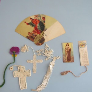 May include: A collection of religious items, including a fan with a religious painting, several crocheted crosses, a bookmark, and a small religious card. The items are arranged on a light blue surface.